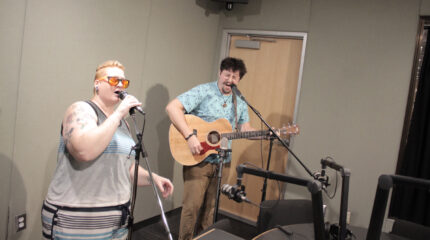 An adult singing into a microphone and an adult singing into a microphone and playing acoustic guitar in a radio studio