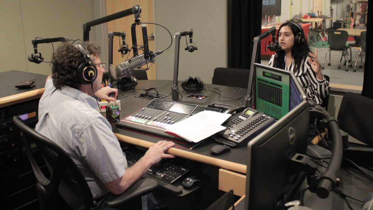 A teen wearing headphones speaking into a microphone and gesturing with their hands being interviewed by an adult wearing headphones sitting behind a microphone and a mixing board in a radio studio