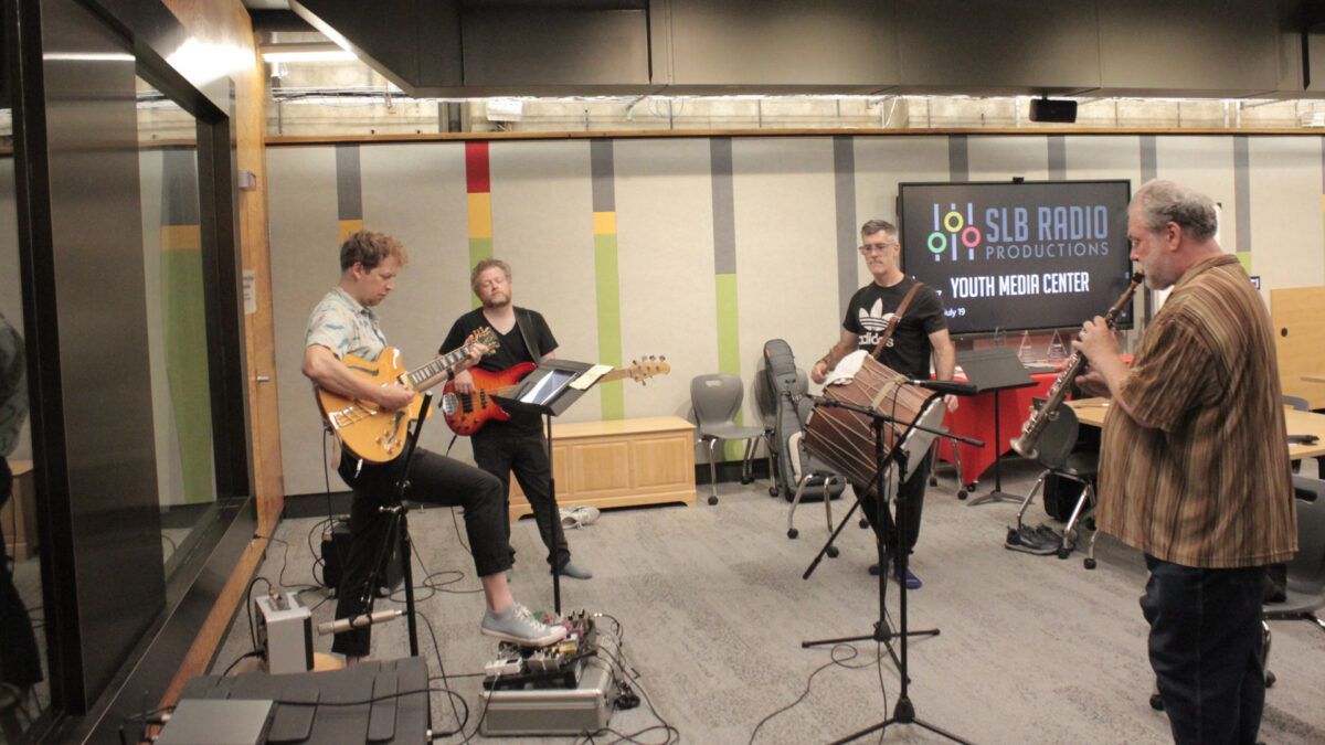 An adult playing electric guitar, an adult playing electric bass, an adult playing tapan, and an adult playing soprano saxophone.
