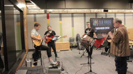 An adult playing electric guitar, an adult playing electric bass, an adult playing tapan, and an adult playing soprano saxophone.