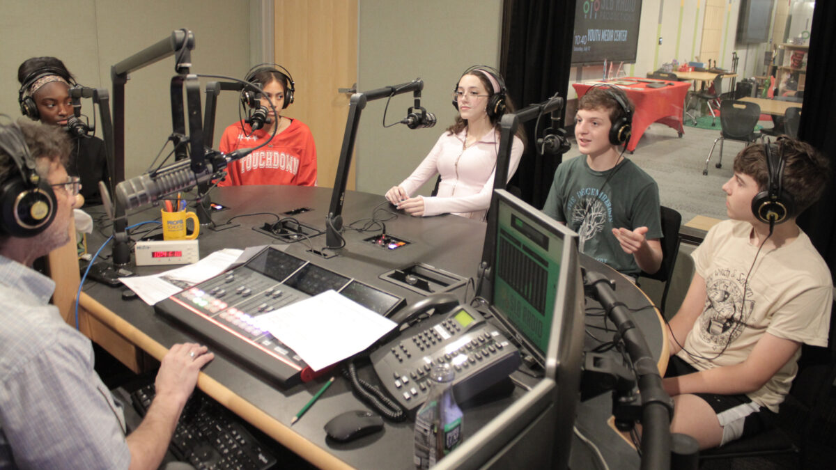 Five youth wearing headphones speaking into microphones being interviewed by an adult wearing headphones sitting behind a microphone and a mixing board in a radio studio