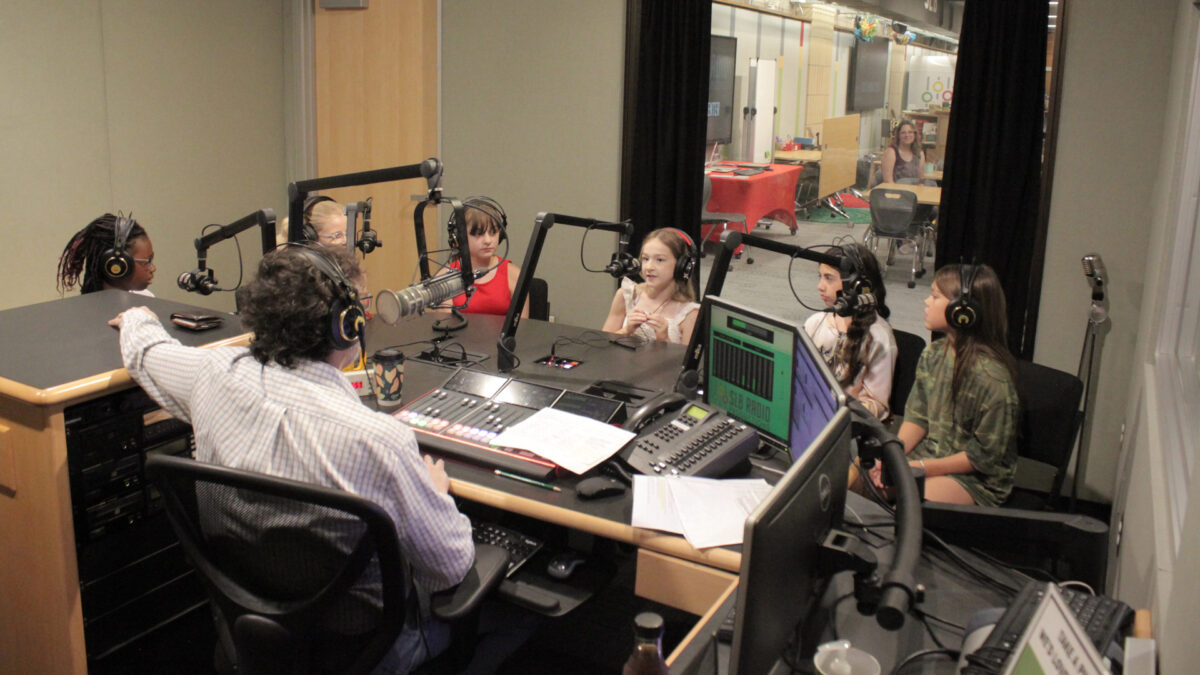 Six youth wearing headphones speaking into microphones being interviewed by an adult wearing headphones sitting behind a microphone and a mixing board in a radio studio