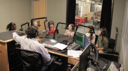 Six youth wearing headphones speaking into microphones being interviewed by an adult wearing headphones sitting behind a microphone and a mixing board in a radio studio