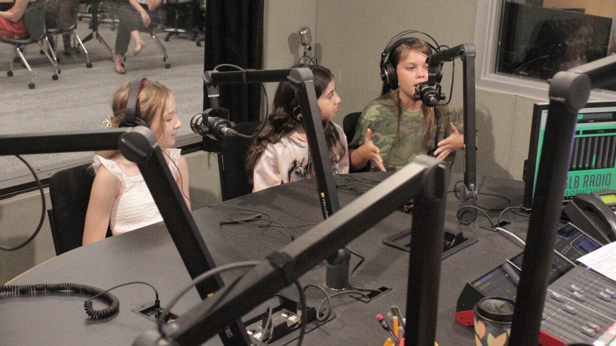 Three youth wearing headphones speaking into microphones, one gesturing with their hands, in a radio studio