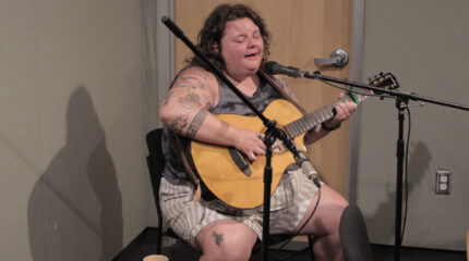 An adult playing acoustic guitar and singing into a microphone in a radio studio