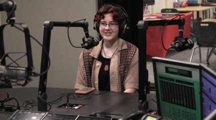A teen wearing headphones smiling behind a microphone in a radio studio