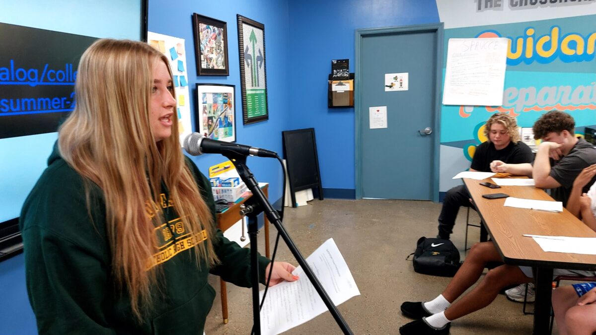 A teen holding a piece of paper and speaking into a microphone as three teens sitting at a long wooden desk listen in a blue classroom