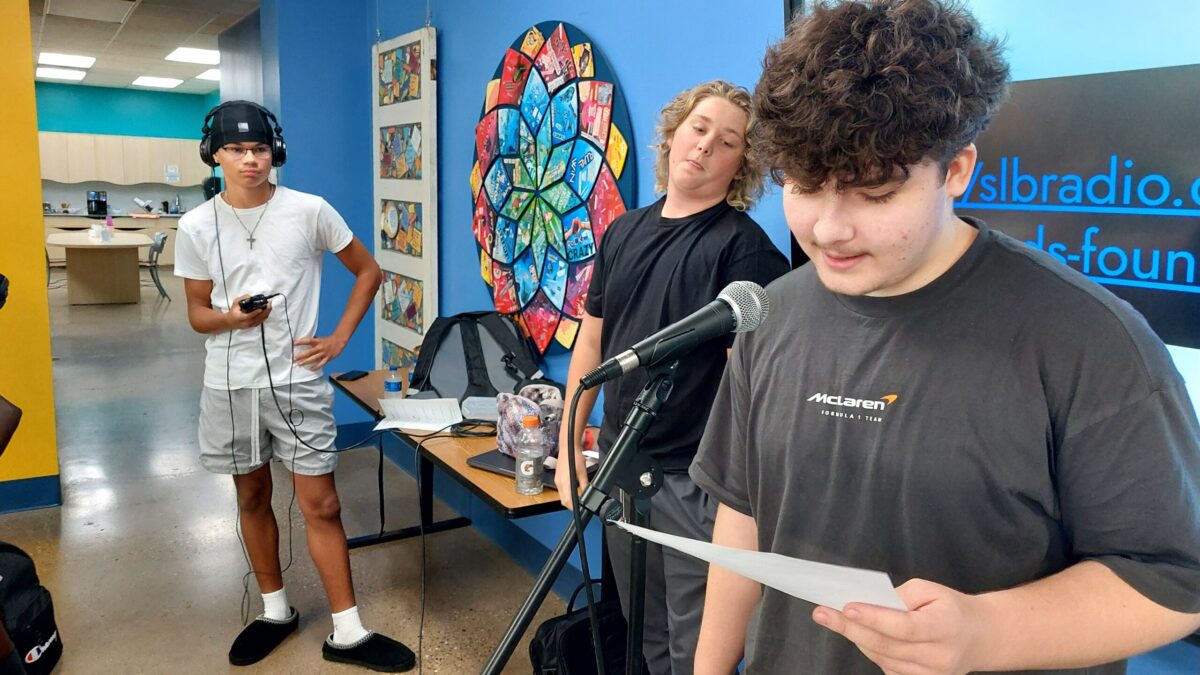 A teen wearing headphones holding a portable recorder and a teen looking at another teen holding a piece of paper and speaking into a microphone