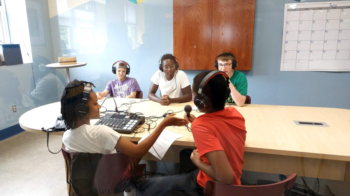 Five teens wearing headphones sitting around a mixer on a wooden table, one holding a microphone