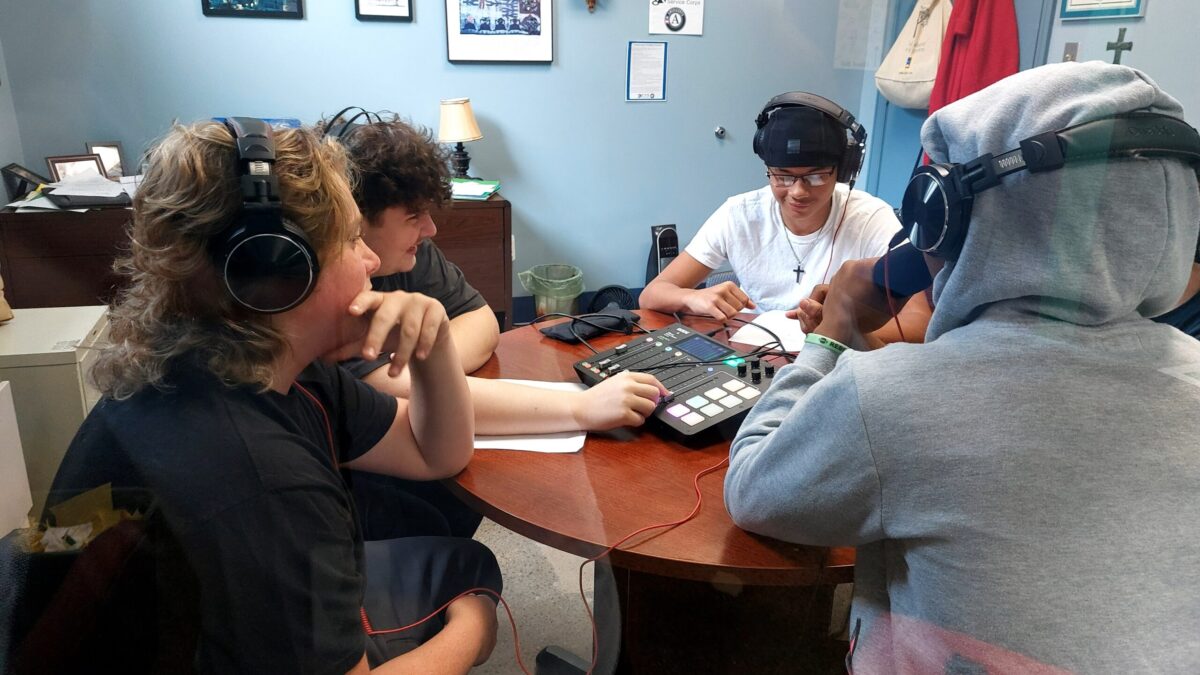 Four teens wearing headphones sitting around a mixer on a round wooden table in a blue office