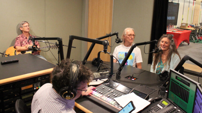 An adult holding an acoustic guitar and two adults speaking into microphones being interviewed by an adult wearing headphones sitting behind a microphone and a mixing board looking at a smartphone in a radio studio