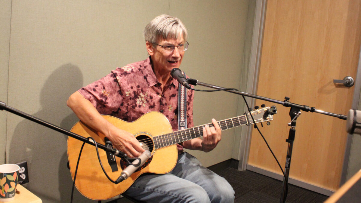 An adult playing acoustic guitar and singing into a microphone in a radio studio