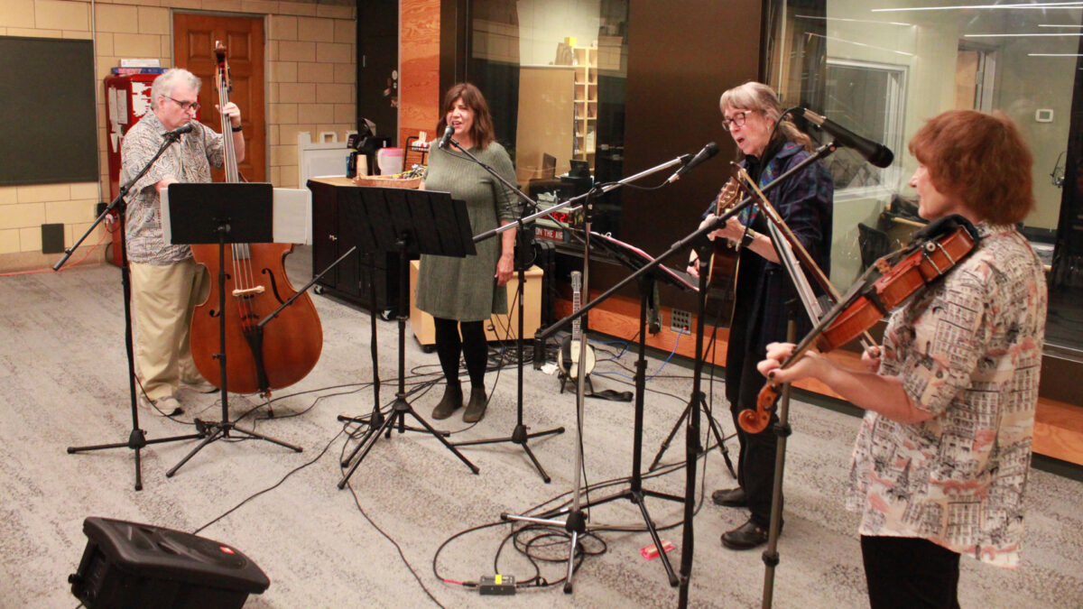 An adult playing upright bass, an adult singing into a microphone, an adult playing acoustic guitar and singing into a microphone, and an adult playing fiddle and singing into a microphone