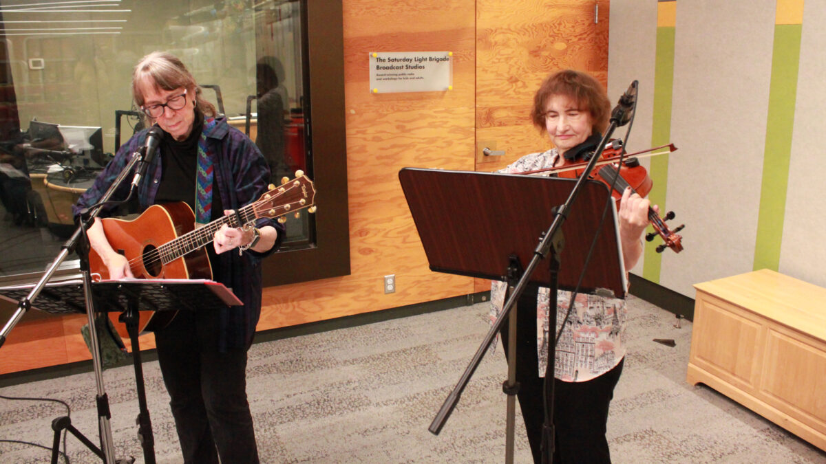 An adult playing acoustic guitar and an adult playing fiddle, both looking at sheet music on music stands