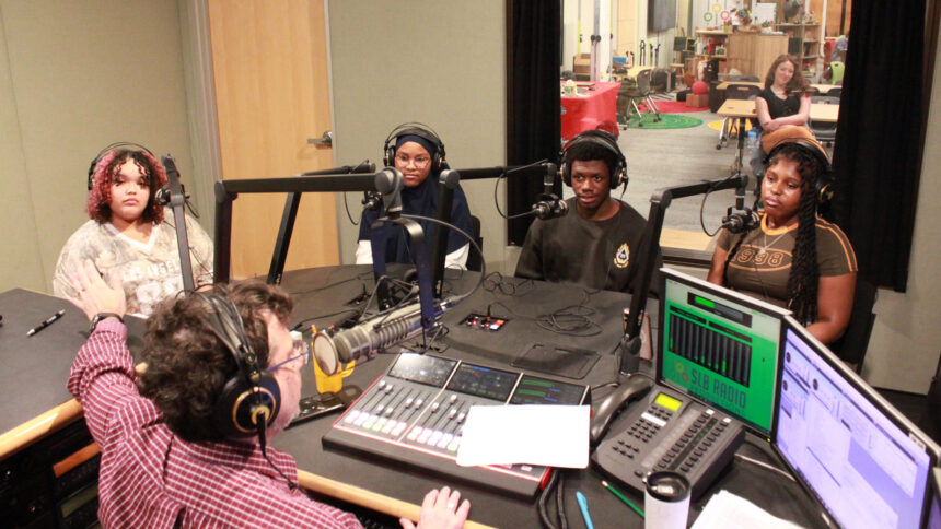 Four teens wearing headphones sitting behind microphones being interviewed by an adult wearing headphones sitting behind a microphone and a mixing board in a radio studio