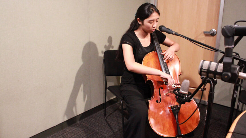 A teen playing a cello sitting behind microphones in a radio studio