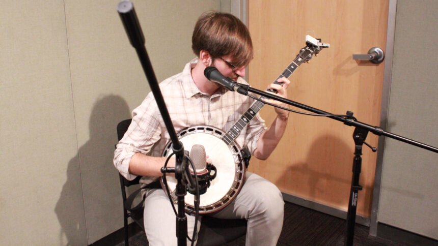 An adult playing banjo sitting behind microphones in a radio studio