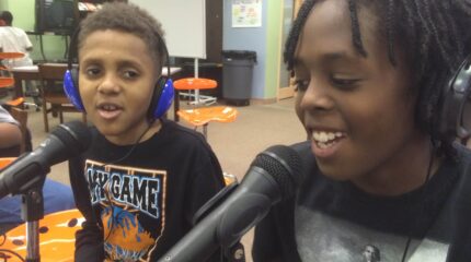 Two youth wearing headphones speaking into microphones in a library