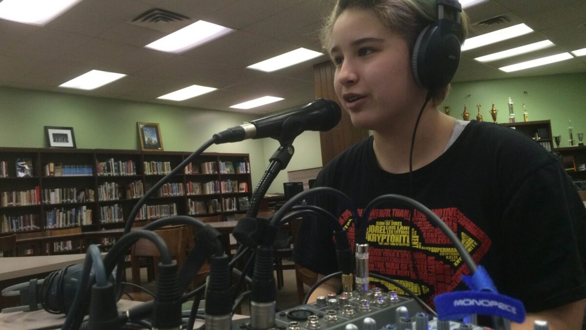 A youth wearing headphones speaking into a microphone connected to a mixer in a library