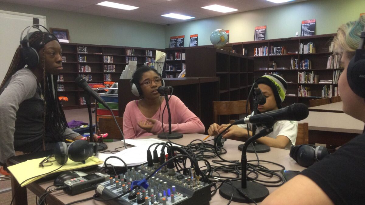 Four youth wearing headphones speaking into microphones connected to a mixer in a library