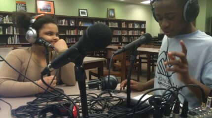 Two youth wearing headphones speaking into microphones connected to a mixer in a library