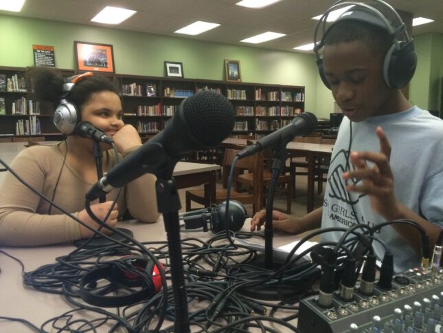 Two youth wearing headphones speaking into microphones connected to a mixer in a library
