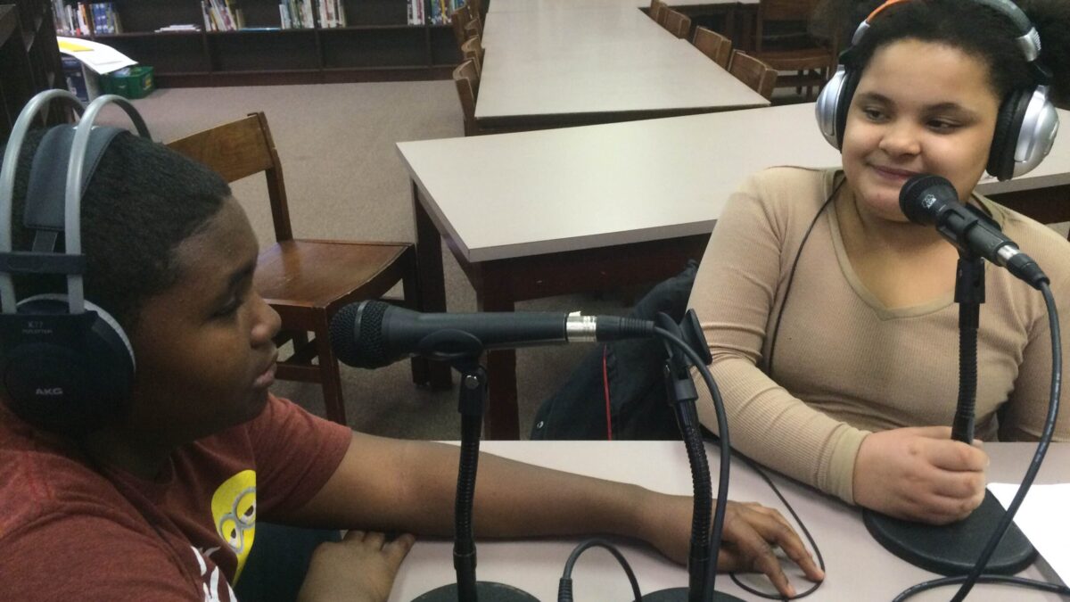 Two youth wearing headphones sitting behind microphones in a library