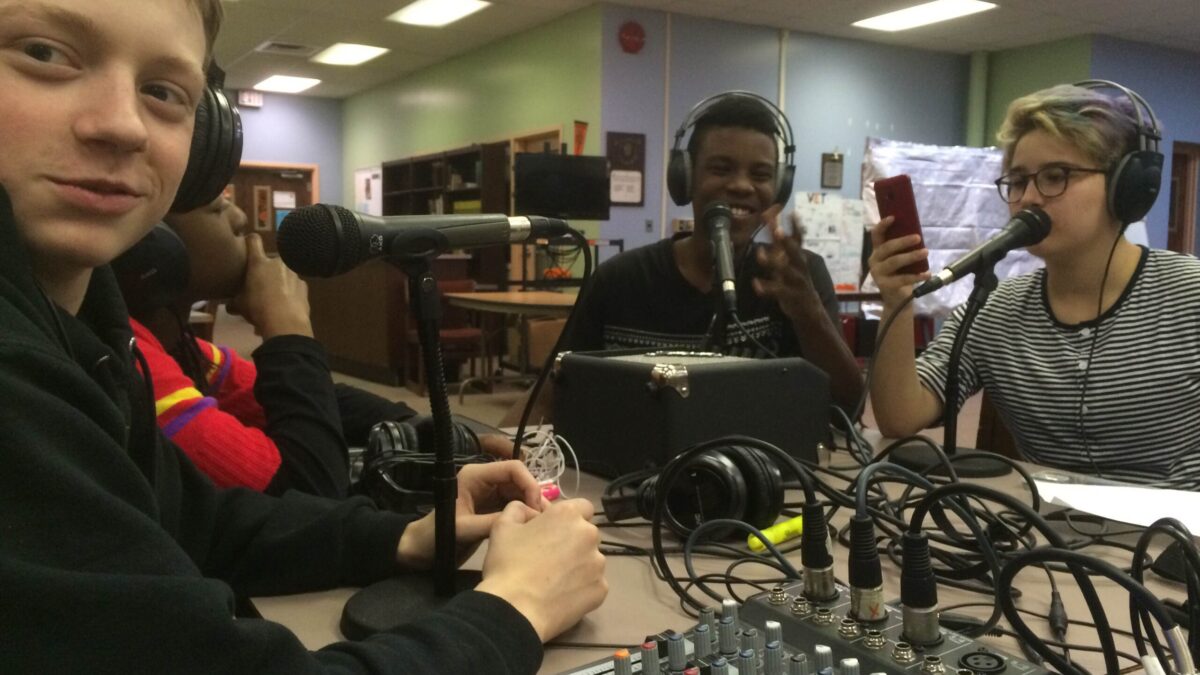 Four youth wearing headphones speaking into microphones connected to a mixer in a library
