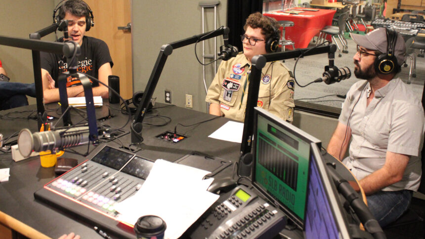 Two adults and a teen wearing headphones speaking into microphones in a radio studio