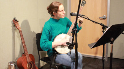 An adult playing a banjo and singing into a microphone in a radio studio