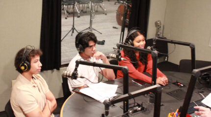 Three teens wearing headphones speaking into microphones being interviewed in a radio studio