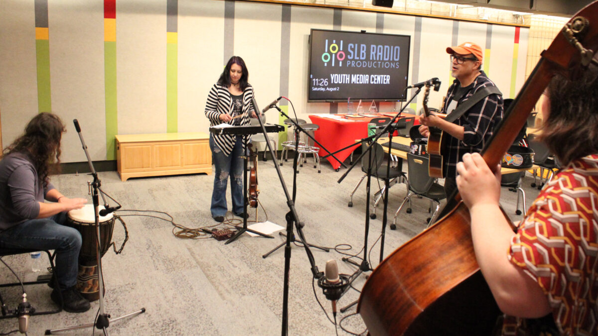 An adult playing djembe, an adult playing glockenspiel, an adult playing acoustic guitar and singing into a microphone, and an adult playing upright bass