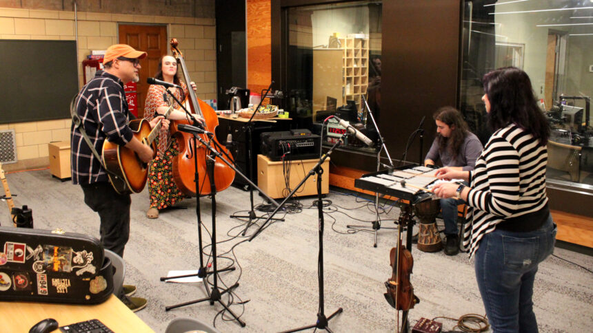 An adult playing acoustic guitar and singing into a microphone, an adult playing upright bass, an adult playing djembe, and an adult playing glockenspiel