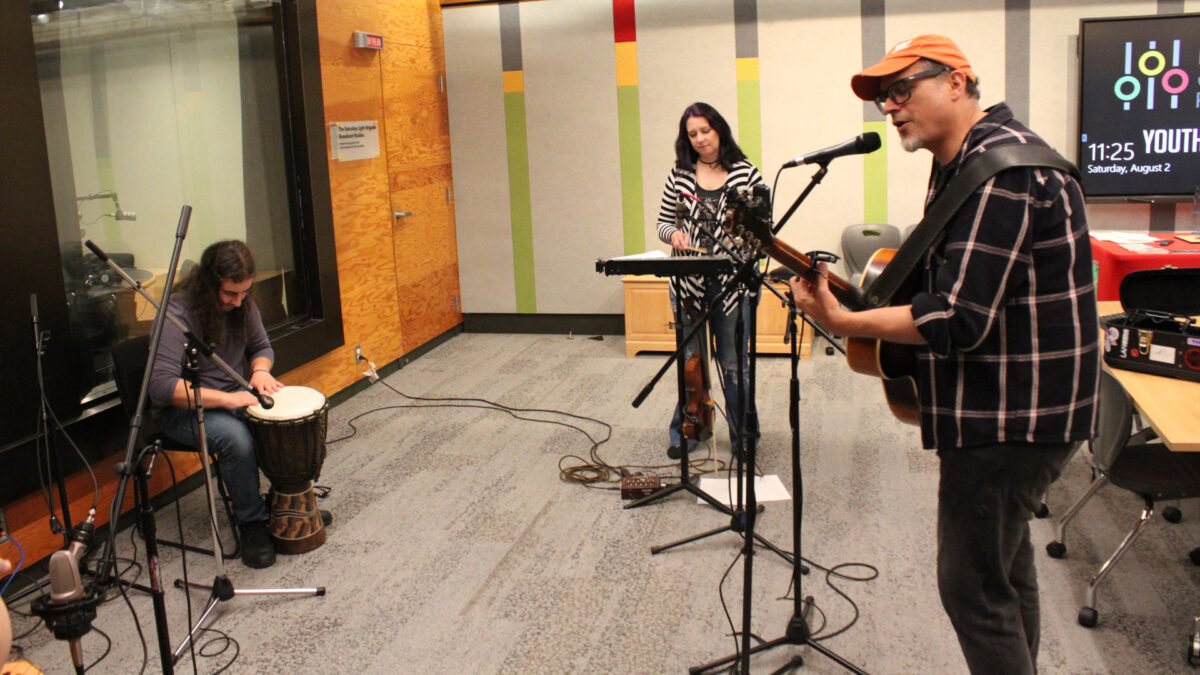 An adult playing djembe, an adult playing glockenspiel, and an adult playing acoustic guitar and singing into a microphone