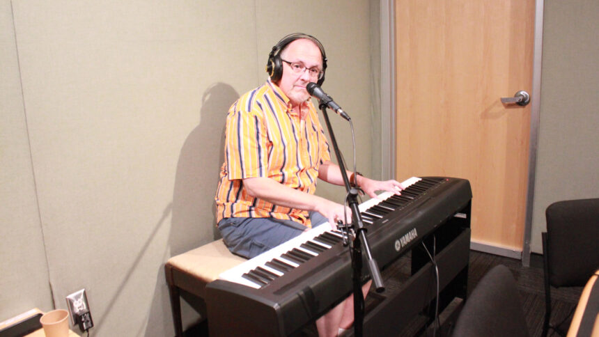 An adult wearing headphones playing a piano keyboard sitting behind a microphone in a radio studio