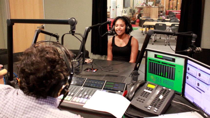 A teen wearing headphones sitting behind a microphone being interviewed by an adult wearing headphones sitting behind a mixing board in a radio studio