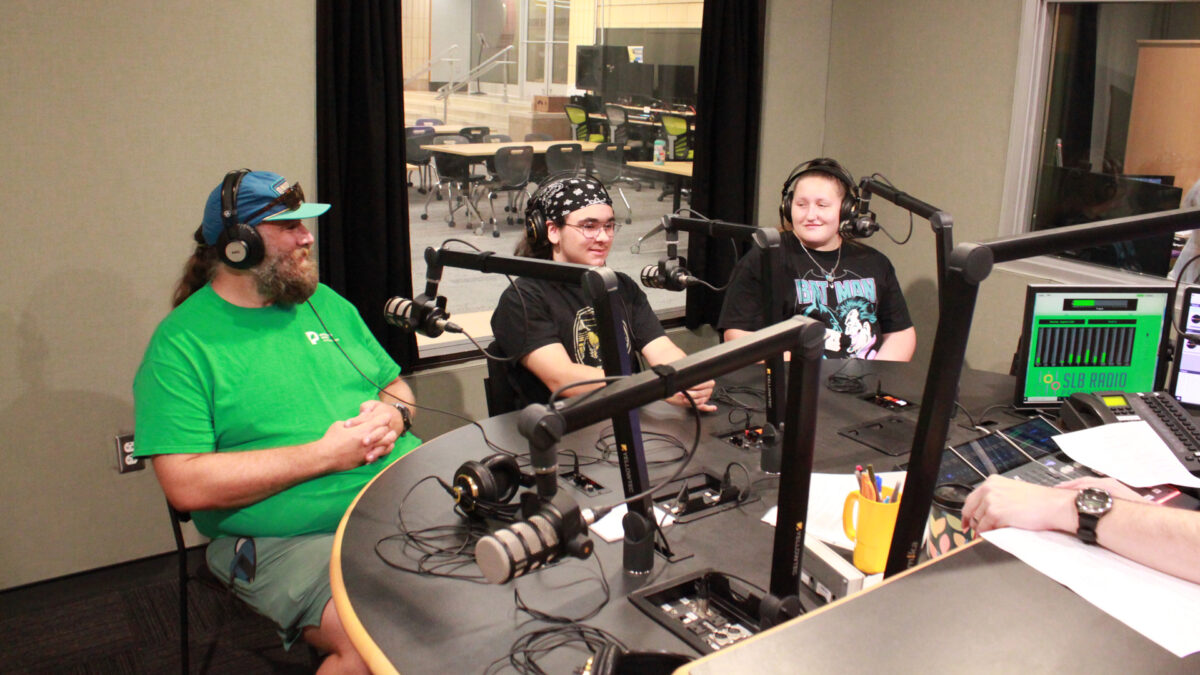 Two teens and an adult wearing headphones sitting behind microphones in a radio studio