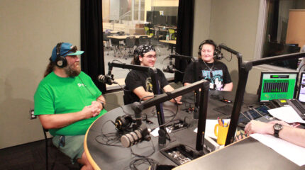 Two teens and an adult wearing headphones sitting behind microphones in a radio studio