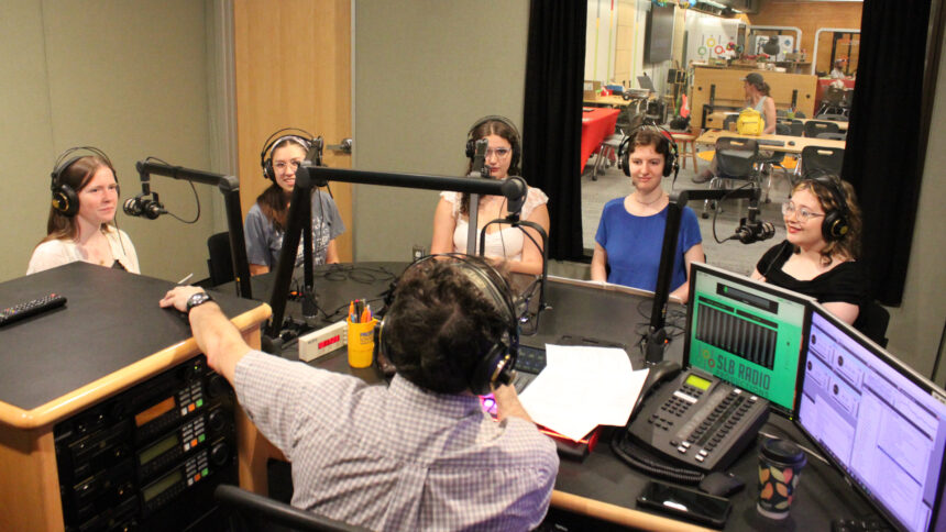 Five teens wearing headphones sitting behind microphones being interviewed by an adult wearing headphones sitting behind a microphone in a radio studio