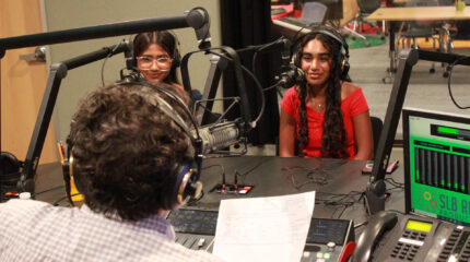 Two teens wearing headphones sitting behind microphones being interviewed by an adult wearing headphones sitting behind a microphone and a mixing board holding a piece of paper in a radio studio