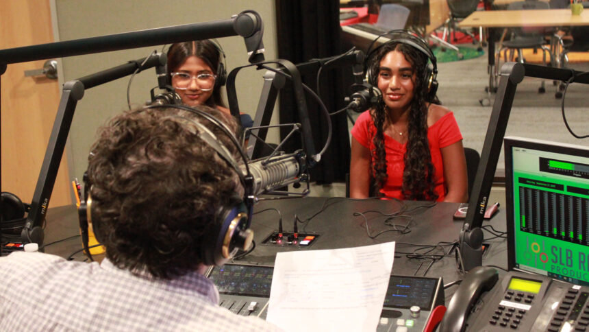 Two teens wearing headphones sitting behind microphones being interviewed by an adult wearing headphones sitting behind a microphone and a mixing board holding a piece of paper in a radio studio