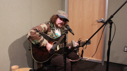 An adult playing an acoustic guitar and singing into a microphone in a radio studio