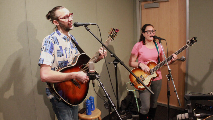 An adult singing into a microphone and playing acoustic guitar and an adult playing electric bass singing into a microphone in a radio studio