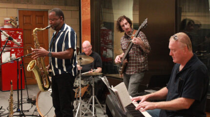 four musicians playing saxophone, drum kit, bass, and keyboard respectively