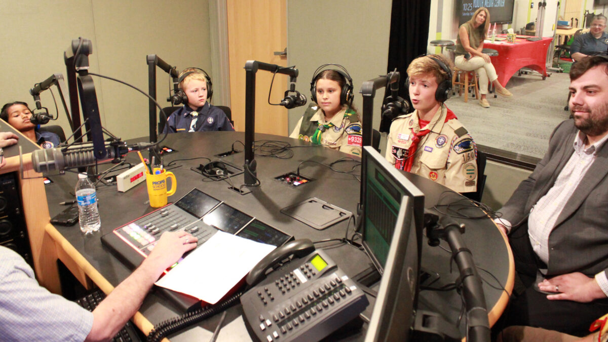 Four youth and one adult wearing headphones speaking into microphones being interviewed by an individual sitting behind a microphone and a mixing board in a radio studio
