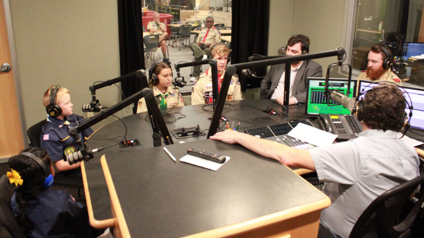 Four youth and two adults wearing headphones speaking into microphones being interviewed by an adult wearing headphones sitting behind a microphone and a mixing board in a radio studio