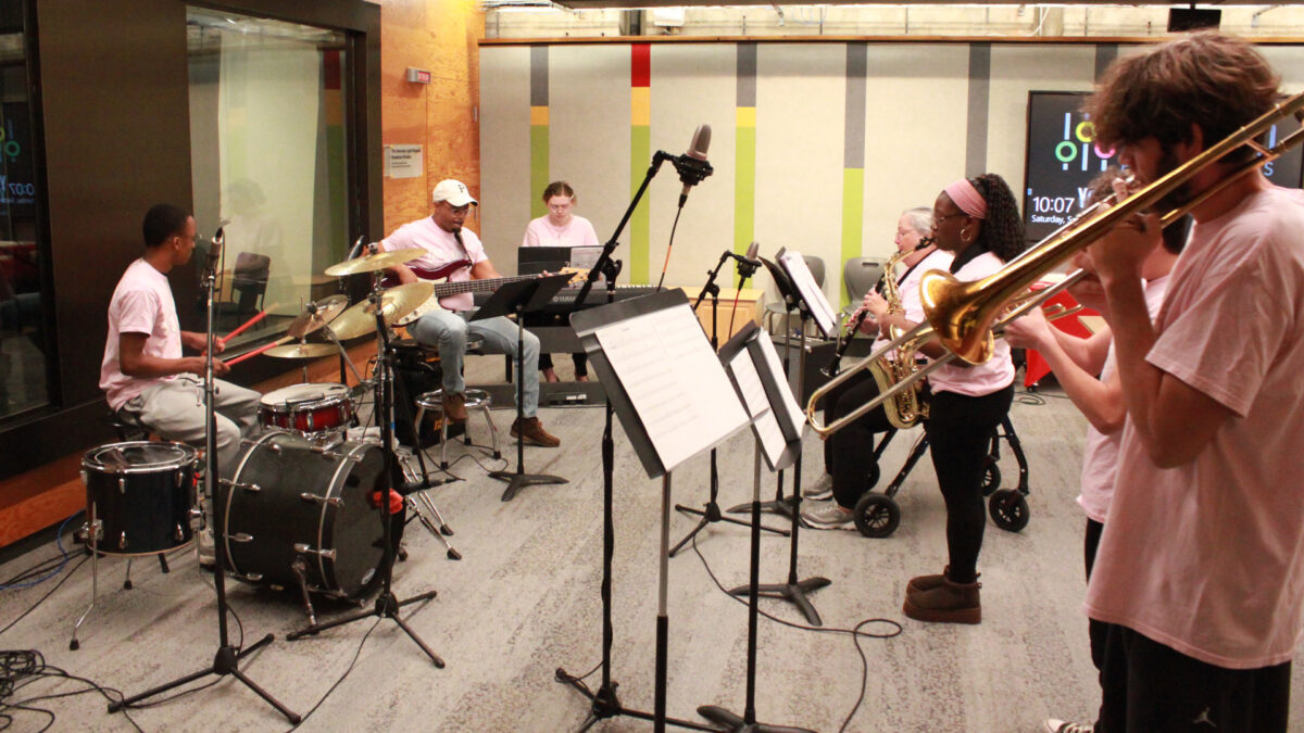 A teen playing a drum set, an adult playing an electric bass, a teen playing a piano keyboard, an adult playing clarinet, a teen playing alto saxophone, a teen playing trumpet, and a teen playing trombone
