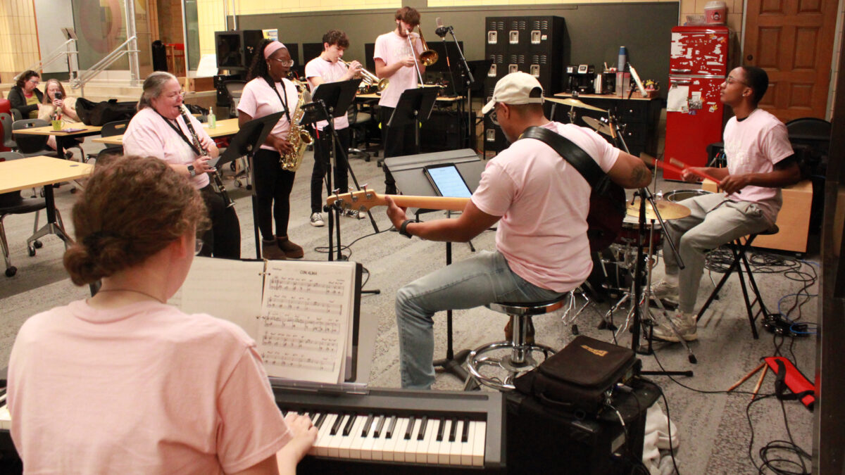 A teen playing a drum set, an adult playing an electric bass, a teen playing a piano keyboard, an adult playing clarinet, a teen playing alto saxophone, a teen playing trumpet, and a teen playing trombone