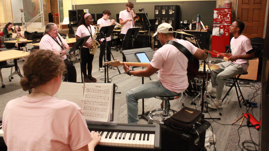 A teen playing a drum set, an adult playing an electric bass, a teen playing a piano keyboard, an adult playing clarinet, a teen playing alto saxophone, a teen playing trumpet, and a teen playing trombone