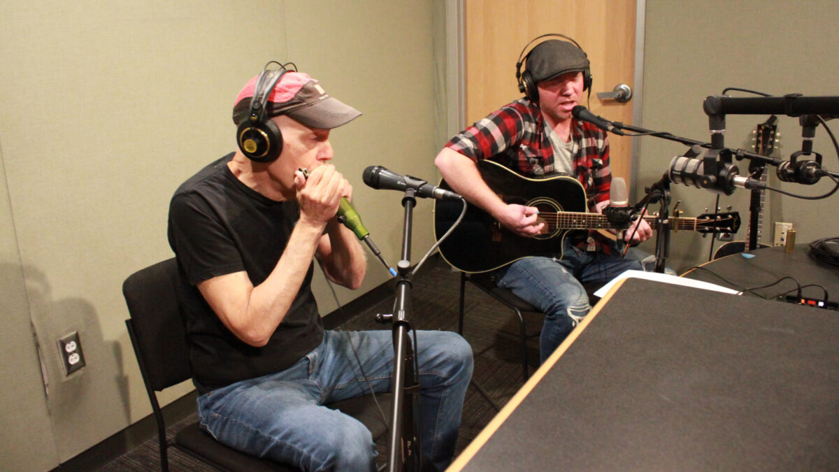 An adult wearing headphones playing harmonica into a microphone and an adult wearing headphones playing an acoustic guitar and singing into a microphone in a radio studio